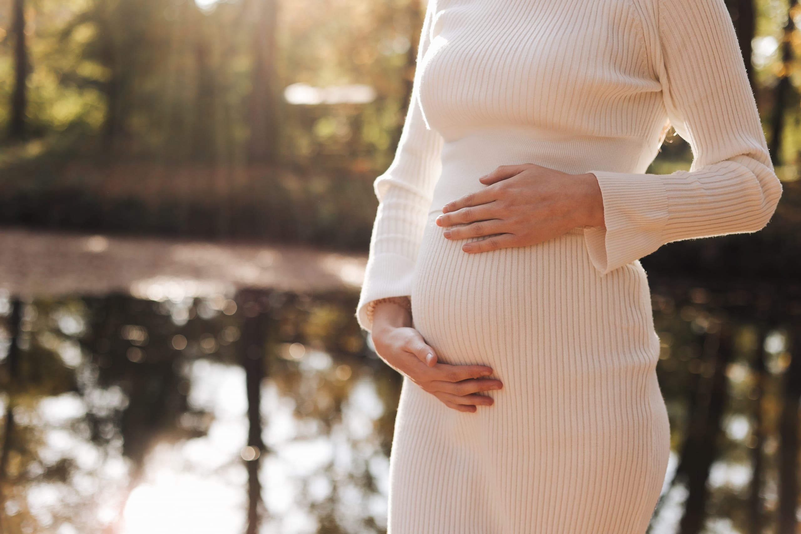 Gravid kvinne i hvit kjole holder hendene rundt magen utendørs