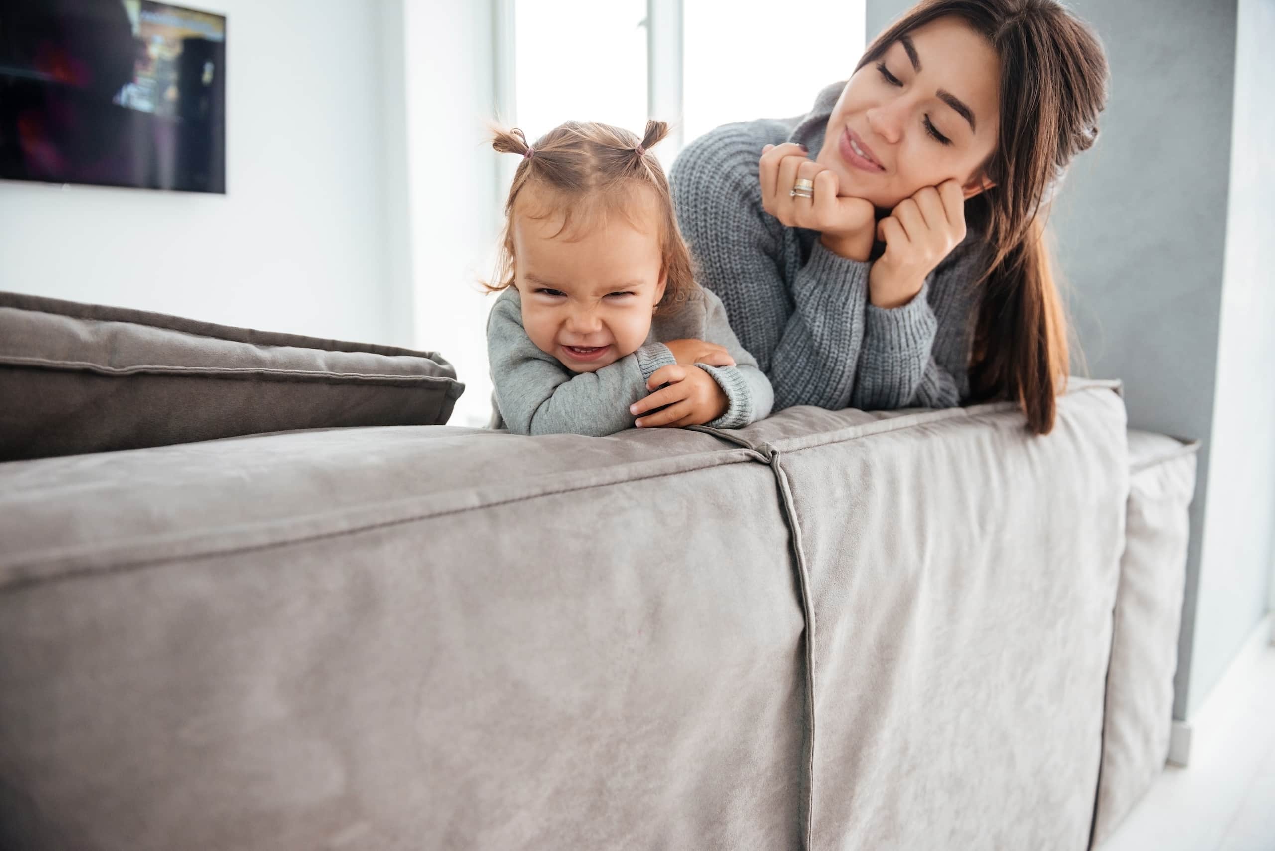 Mor og lite barn lener seg over ryggen på en sofa